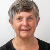 A woman with short gray hair smiles warmly at the camera, wearing a black top and delicate jewelry, against a simple white background.