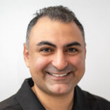 A smiling man with dark hair, wearing a black shirt, is looking directly at the camera. His friendly expression conveys warmth and approachability, making the image inviting.