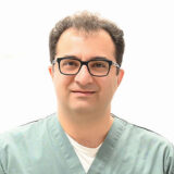 A dental professional wearing scrubs and glasses smiles at the camera against a bright background, exuding a welcoming demeanor and a confident presence.