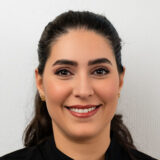 A smiling woman with long dark hair is wearing a black top against a light background. She has a friendly expression, showcasing her bright smile and subtle makeup, suggesting a welcoming demeanor.