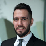 A man in a dark suit and tie smiles confidently, showcasing a well-groomed beard and short hair. He stands with a modern, softly blurred background, projecting professionalism and approachability.