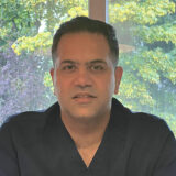 A man wearing a dark shirt is sitting in front of a window with lush greenery outside. He has a neutral expression and appears to be in a professional setting.