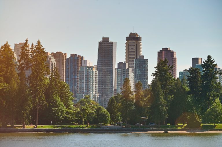 A city skyline with tall skyscrapers and lush green trees by a calm water body, featuring a large blue smiling face in the sky.