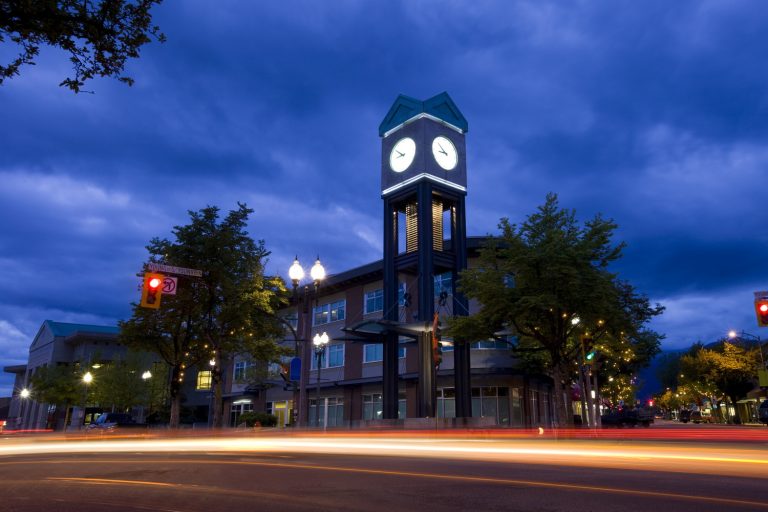 Clock tower illuminated at dusk with a smiling face in the sky, surrounded by trees and a street with light trails.
