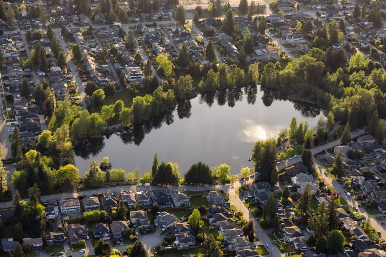 Aerial view of a suburban neighborhood with a central lake surrounded by houses and trees, under a sunlit sky.