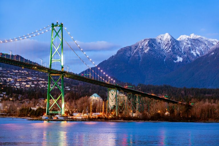 A colorful suspension bridge spans a river, with snow-capped mountains in the background. A cheerful, smiling icon overlays the scene, adding a vibrant touch to the landscape at dusk.