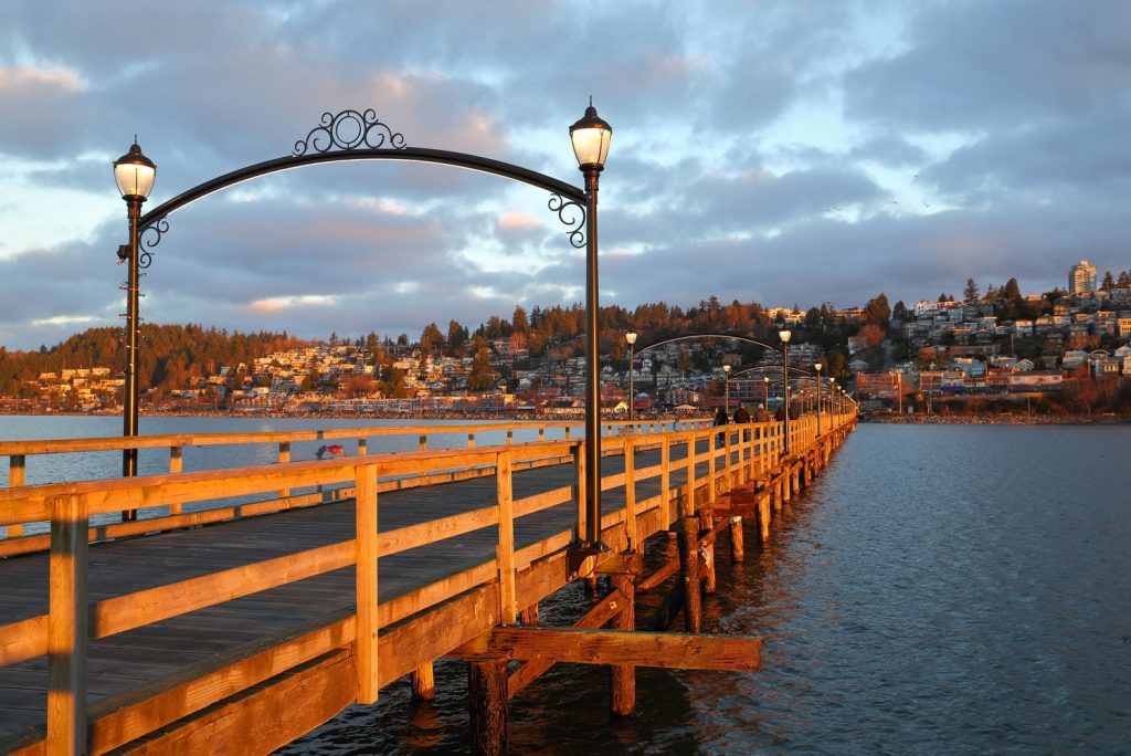 A long wooden pier stretches into the distance over calm waters, with a decorative arch and lanterns. Behind it, a scenic view of a town at sunset, suggesting a peaceful locale. Perfect spot to explore after visiting a dentist near you for a relaxing stroll.