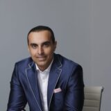A professional man in a tailored blue suit is seated at a desk, smiling confidently. He has short dark hair and is wearing a white shirt with a pink pocket square, set against a neutral background.