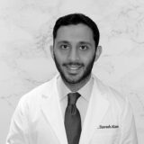 A smiling male doctor wearing a white coat and tie stands in front of a marble background. His name, Slavash Alam, is visible on the coat.