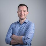 A smiling man with short hair stands confidently with his arms crossed, wearing a light blue shirt against a plain gray background.