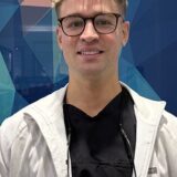 A young man with blonde, wavy hair and glasses smiles at the camera, wearing a black shirt and a light jacket. The background features abstract geometric patterns in various colors.