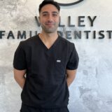 A smiling male dentist stands in front of a "Valley Family Dentist" sign, wearing black scrubs. The background features a textured wall, emphasizing a professional and welcoming dental environment.
