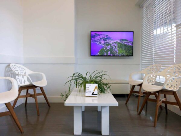 A modern waiting room featuring white, decorative chairs, a sleek coffee table, and a potted plant. A television displays a vibrant image of a coastal landscape. Natural light filters through blinds, creating a bright and inviting atmosphere.