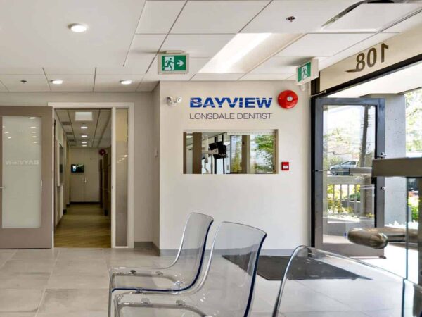 A modern dental clinic reception area featuring a sign for "Bayview Dental," transparent chairs, and a large entrance door. The space is well-lit with neutral-colored walls and a visible hallway leading to treatment rooms.