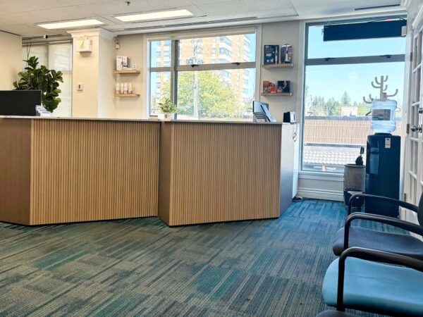 A bright, modern reception area featuring a wooden front desk, green carpet, and seating. Large windows provide natural light, and a potted plant adds a touch of warmth to the space.