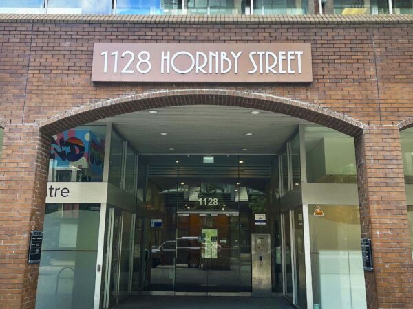 The image shows the entrance of a building at 1128 Hornby Street, featuring a prominent sign above the doorway. The structure has a modern design with glass panels and a brick façade.