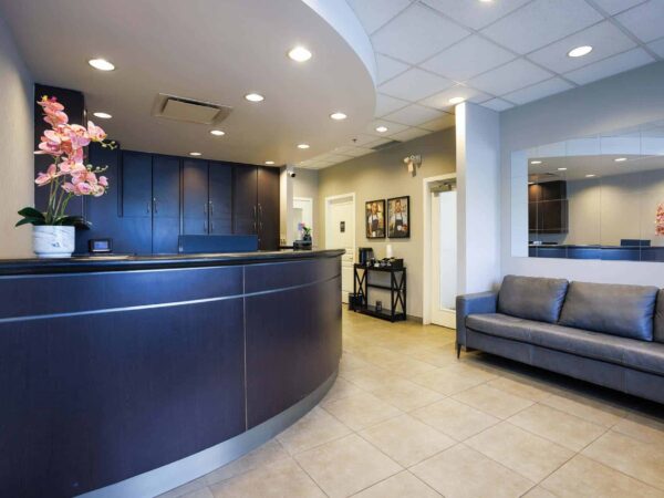 A modern reception area featuring a curved dark wooden desk, a potted pink flower arrangement, a gray couch, and framed pictures on the wall. The space is well-lit with a clean, inviting atmosphere.