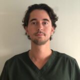 A young man with curly dark hair is wearing a green medical scrub top. He stands against a plain background, looking directly at the camera with a neutral expression.