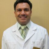 A smiling man wearing a white lab coat and a green striped tie stands against a beige background, presenting a professional and approachable demeanor.