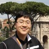 A person with glasses and short hair smiles in front of a historic archway and lush green trees, suggesting a sunny day and a visit to a notable site.