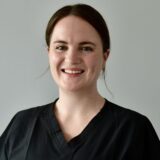 A smiling female healthcare professional wearing a black uniform. She has shoulder-length brown hair and is facing the camera against a neutral background.