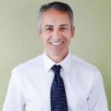 A smiling man dressed in a white shirt and patterned tie stands against a light green background. He has short, neatly styled hair and appears friendly and approachable.