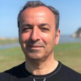 A smiling man with short, dark hair stands outdoors by a body of water, wearing a black shirt. The background features a grassy area and hints of a shoreline under a clear blue sky.