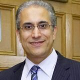 A man with gray hair and glasses smiles, wearing a dark suit and light-colored shirt with a tie. The background features wooden paneling, suggesting a formal setting.