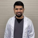 A smiling individual wearing a white lab coat over gray scrubs stands against a neutral background, conveying a professional healthcare or medical setting.