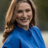 A woman with wavy brown hair smiles warmly while wearing a blue top. The background features soft, natural greenery, creating a friendly and inviting atmosphere.