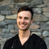 A smiling young man wearing a black shirt stands confidently in front of a stone wall, suggesting a friendly and approachable demeanor.