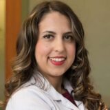 A smiling woman with long, wavy brown hair poses in a professional white coat. She stands against a soft-focus background, exuding confidence and approachability.