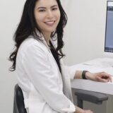 A woman with long dark hair, wearing a white lab coat, is sitting in front of a computer in a modern office setting, smiling at the camera.