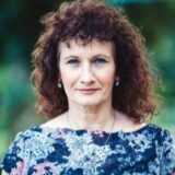A woman with curly hair and blue eyes is outdoors, wearing a floral dress. She has a confident expression and is looking directly at the camera.