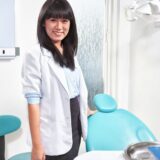 A smiling female dentist stands beside a dental chair in a bright clinic. She is wearing a white coat over a blue shirt and a black skirt, with dental equipment visible nearby.
