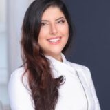 A smiling woman with long, dark hair in a white jacket. She stands against a softly blurred background, exuding a warm and friendly demeanor.