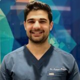 A smiling male healthcare professional wearing a dark scrubs top stands in front of a colorful, abstract background. His name, Dr. Arian Majid, is embroidered on his scrubs.
