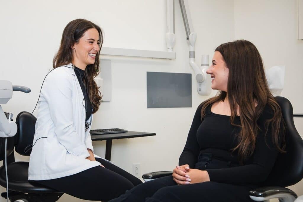 A smiling dentist in a white coat and a patient in a black outfit share a friendly conversation while seated in a dental office. The atmosphere is relaxed and welcoming, highlighting a positive patient experience.