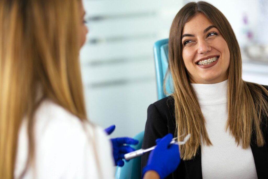 A smiling woman in a dental chair engages with a dentist, who is wearing gloves and holding a dental tool. The atmosphere seems friendly and positive, reflecting a comfortable dental visit experience.