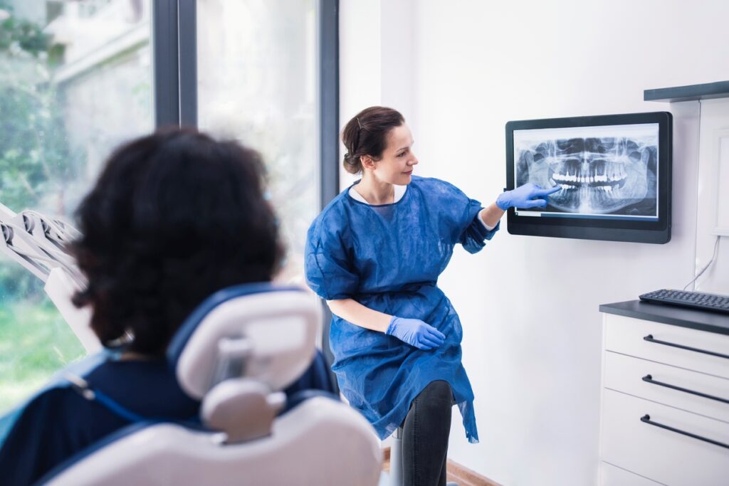 A dental professional in a blue gown explains an X-ray displayed on a screen to a patient seated in a dental chair. The environment is bright and modern, suggesting a calm atmosphere for dental care.