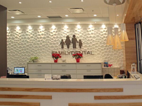 A modern dental clinic reception area featuring a white textured wall, the words "Family Dental," decorative plants, and a clean, organized front desk with computer screens and brochures. Soft lighting enhances the welcoming atmosphere.