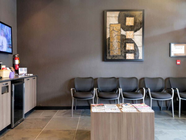 A modern waiting room featuring gray walls, several black chairs, a coffee station, a flat-screen TV displaying content, and a wooden table with magazines. An abstract artwork is hung on the wall, contributing to the contemporary aesthetic of the space.