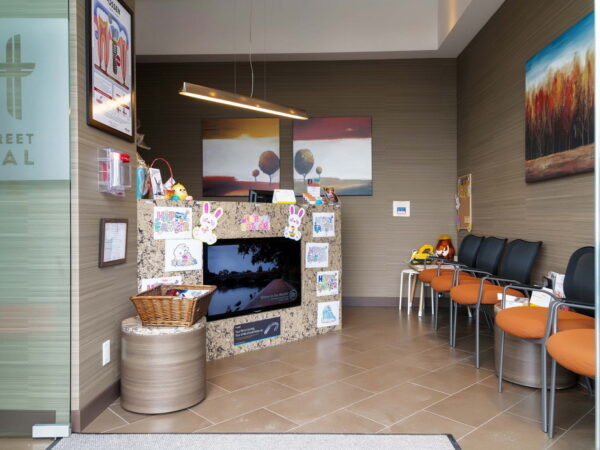 A dental office waiting area featuring orange chairs, a decorative wall with kids' artwork, and contemporary art pieces. A cozy, inviting atmosphere with natural light and subtle decor elements.