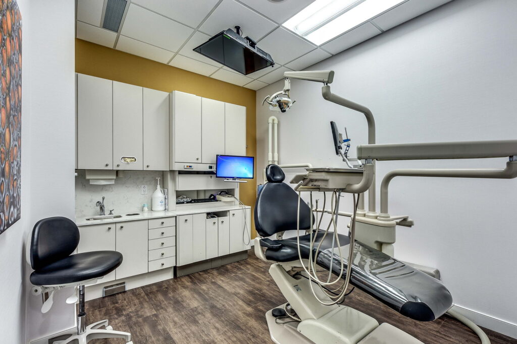 A modern dental office featuring a dental chair with overhead lighting, a computer screen, and storage cabinets against a wall. The room has a neutral color scheme with wooden flooring and a minimalist design.