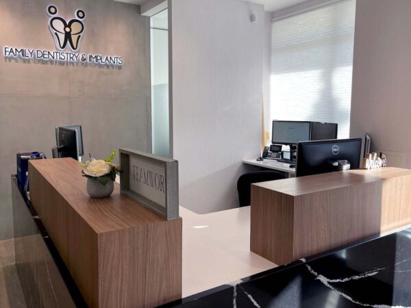 A modern reception area featuring a wooden front desk, two computer stations, and a decorative flower arrangement. The wall displays the name "Family Wellness & Health," indicating the purpose of the space. Natural light comes from large windows, creating a welcoming atmosphere.