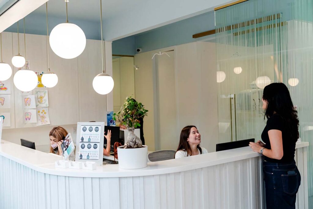 A modern reception area features two staff members at a curved desk with decorative plants and round pendant lights. One staff member interacts with a visitor while another works on a computer, creating a welcoming atmosphere.