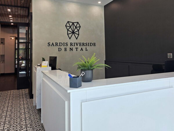 A modern dental office reception area featuring a white desk, the "Sardis Riverside Dental" logo on the wall, and a small indoor plant. The decor includes patterned flooring and contemporary lighting, creating a welcoming atmosphere for patients.