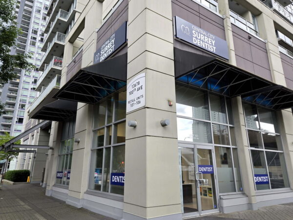Exterior view of a commercial building featuring large windows and an awning, displaying "DENTAL" signage. The structure is part of a modern urban setting with nearby high-rise buildings.