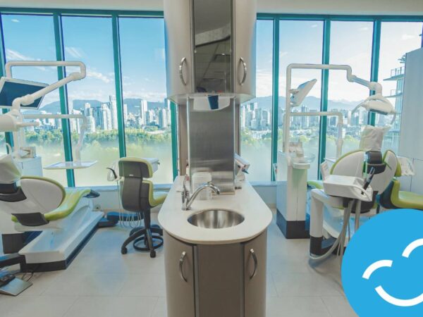 A modern dental clinic with large windows offering a city view. The image features dental chairs, equipment, and a central workstation with a sink, creating a clean and inviting environment for patients.