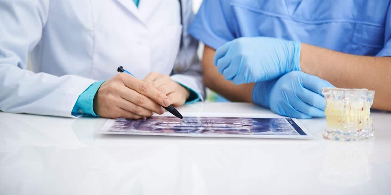 A dental professional in a white coat reviews an X-ray with a colleague wearing blue gloves, with a glass of water nearby. The setting suggests a clinical environment focused on dental care and patient treatment.
